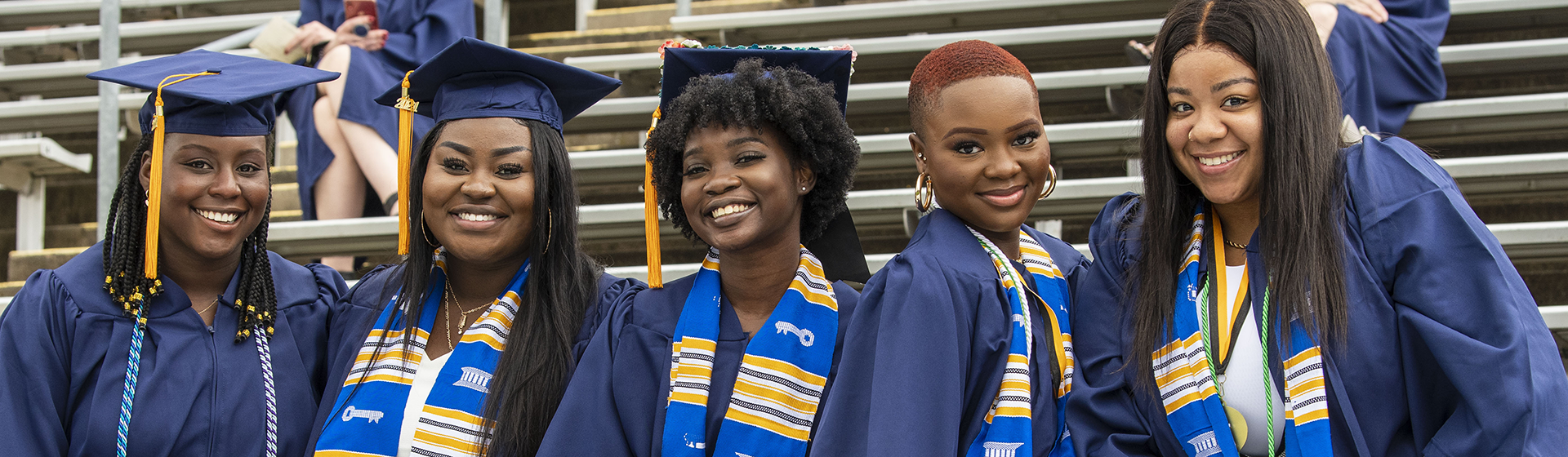 Commencement - 5 students in cap and gown smiling and sitting on bleachers